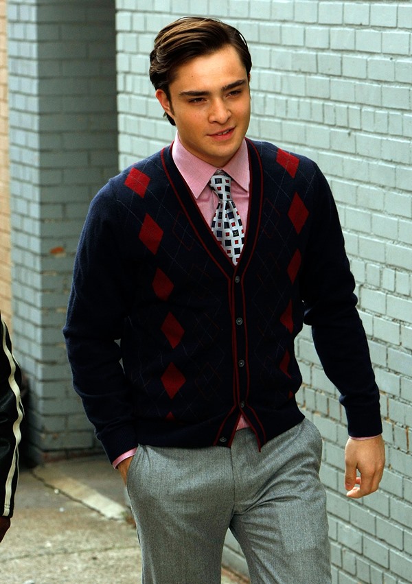 O ator Ed Westwick, um homem branco, jovem, de cabelo liso castanho, durante as gravações da série Gossip Girl em 2008. Ele usa uma camisa rosa de botao, uma gravata branca e um casaco estilo suéter azul