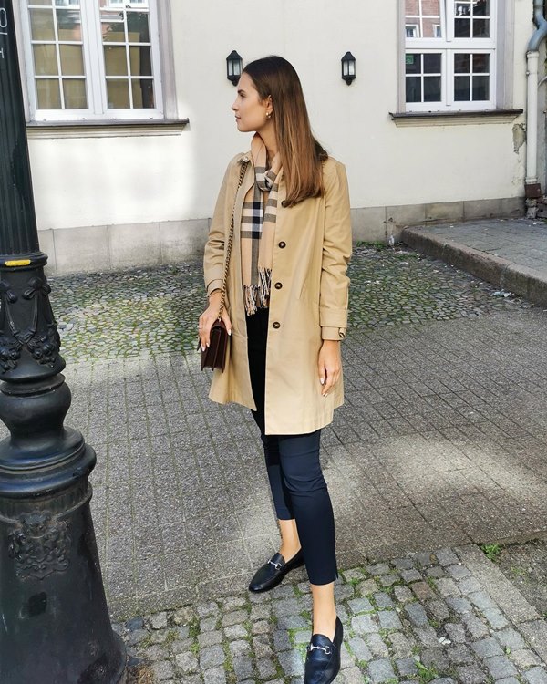 Mulher jovem e branca, de cabelo longo liso e loiro, posando para foto na rua. Ela usa calça jeans, sapatos mocassins, sobretudo bege e um cachecol listrado