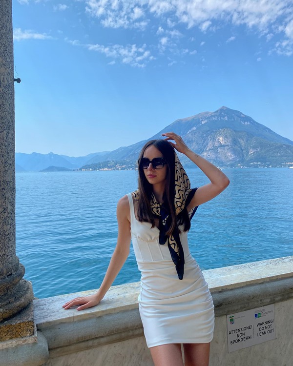 Mulher jovem e branca, de cabelo liso e castanho claro, posando em região do litoral europeu. Ela usa um vestido branco justo, óculos escuros e um lenço de seda na cabeça