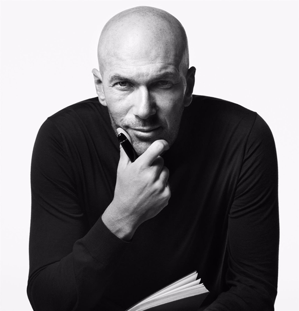 Campanha de divulgação da marca de luxo Montblanc. A foto, feita em preto e branco, mostra o jogador de futebol Zinédine Zidane, um homem branco, de meia idade e careca, com objetos da marca alemã. Ele mostra itens como canetas, pastas e cadernos.