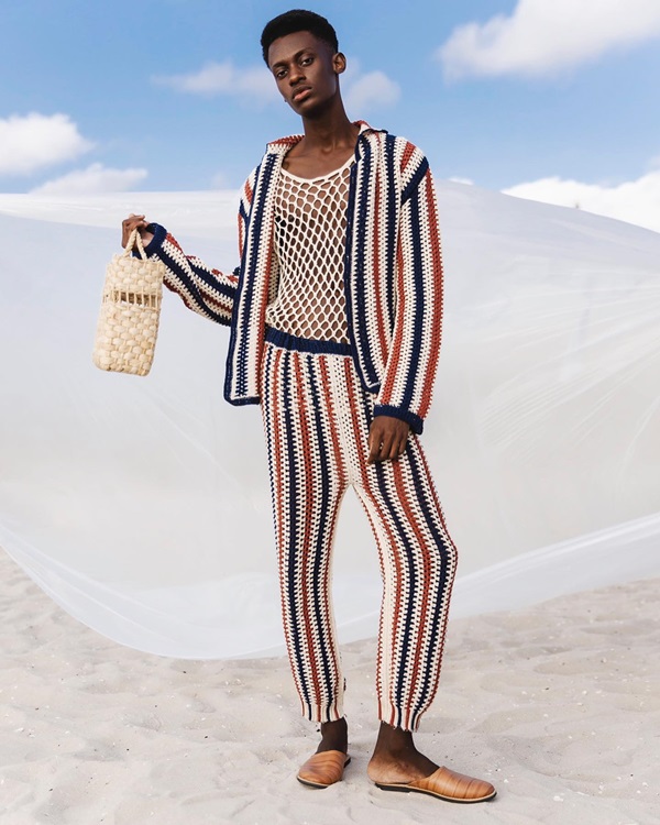 Campanha de divulgação da coleção de roupas da marca Ateliê Mão de Mãe. O modelo, um homem jovem e negro de cabelo raspado, está em uma praia de areia branca e com o céu azul. Veste um conjunto de crochê listrado preto, azul e vermelho e segura uma bolsa de palha em cor cru.