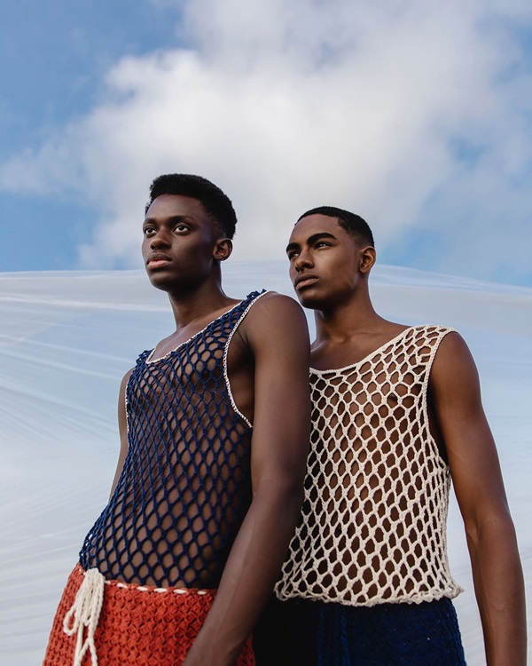 Campanha de divulgação da coleção de roupas da marca Ateliê Mão de Mãe. Os modelos, dois homens jovens e negros, estão em uma praia de areia branca e com o céu azul. Vestem regatas de crochê com pontos bem abertos nas cores azul marinho e branco.