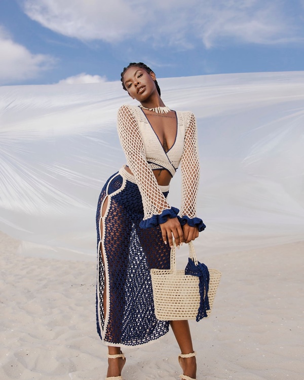 Campanha de divulgação da coleção de roupas da marca Ateliê Mão de Mãe. A modelo, uma mulher jovem e negra de cabelo trançado, está em uma praia de areia branca e com o céu azul. Veste um conjunto de crochê de top e saia longa, ambos nas cores brancas e azul marinho, e segura uma bolsa de palha na cor cru.