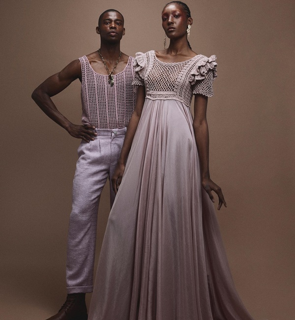 Modelos jovens e negros, um homem e uma mulher, posando para foto em um estúdio marrom. Ele usa uma blusa regata e uma calça e ela um vestido longo. Todas as peças foram confeccionadas em crochê rosa claro. A foto é de campanha da marca Ateliê Mão de Mãe.