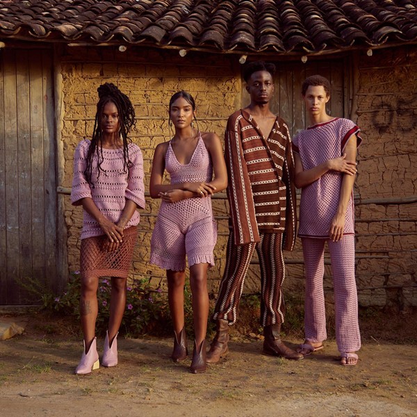 Modelos negros, dois homens e mulheres, posando para foto em um casa feita de barro na Bahia. Eles roupas confeccionadas em crochê marrom em nas cores rosas e marrom. A foto é de campanha da marca Ateliê Mão de Mãe.