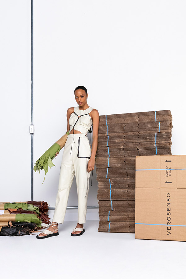 Campanha de divulgação da coleção de verão Angiô da marca Verosenso. Na foto, feita em um fundo branco, é possível ver várias caixas de papelão empilhadas servindo de cenário e apoio para a modelo. Ela, uma mulher jovem negra, de cabelo cacheado amarrado em um coque, usa um conjunto de cropped e calça, ambas peças feitas em couro branco e com detalhes de corda preta.