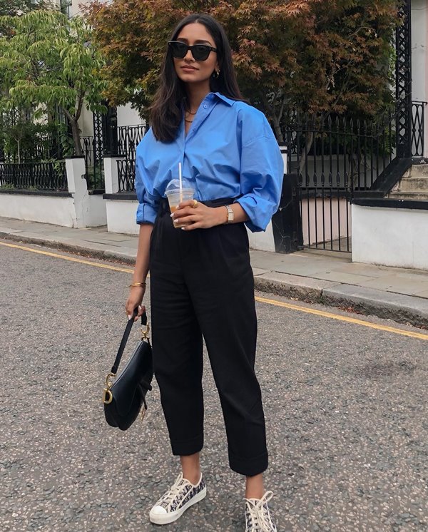 Mulher jovem e de pele morena, com cabelos lisos longos, posando para foto nas ruas de Paris. Ela usa um óculos escuro, uma camisa de botão azul, uma calça preta de alfaiataria, um tênis branco e uma bolsa de couro preta