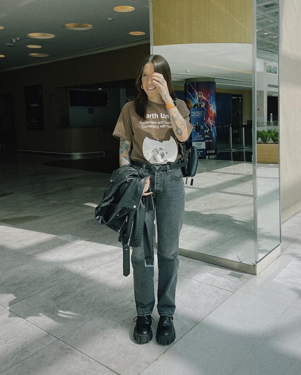 Mulher jovem e branca,, com cabelos lisos longos, posando para foto no corredor de um shopping. Ela usa uma camiseta verde, uma calça jeans cinza, jaqueta de couro, sapato estilo mocassim preto de couro e uma bolsa, também de couro preto.