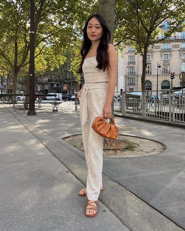 Mulher jovem e de pele clara, com cabelos lisos longos e traços de origem asiática, posando para foto nas ruas de Paris. Ela usa um conjunto de calça e top de linho cru, uma sandália rasteira e uma bolsa pequena, ambas as peças de couro marrom