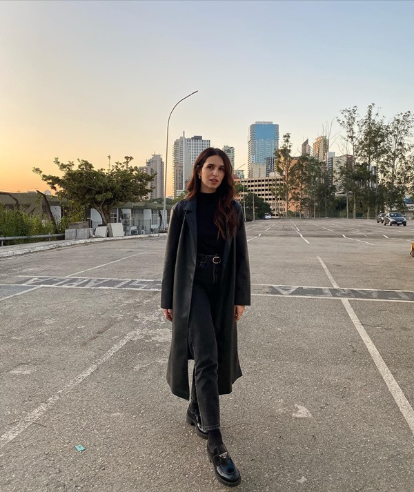 Mulher jovem e branca, com cabelos lisos longos, posando para foto em um estacionamento ao ar livre. Ela usa uma camiseta preta, calça jeans cinza e um casaco estilo sobretudo em crochê preto.