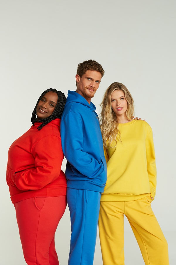 Campanha de divulgação da Simples, submarca da Reserva, onde modelos posam com as roupas em um fundo branco. Na foto, é possível ver três modelos usando moletons da marca nas cores azul, amarelo e vermelho. 