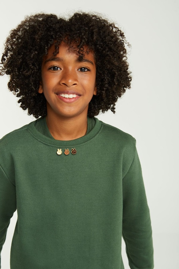 Campanha de divulgação da Simples, submarca da Reserva, onde modelos posam com as roupas em um fundo branco. Na foto, um menino negro de no máximo 7 anos com cabelo cacheado estilo black power e um moletom verde escuro.