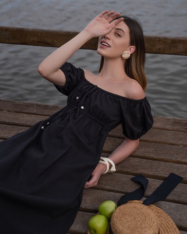 Mulher branca e jovem, de cabelo liso loiro, posando para foto em um deck de mandeira na beira do lago. Ela usa um vestido preto de mangas bufantes e decote estilo ciganinha