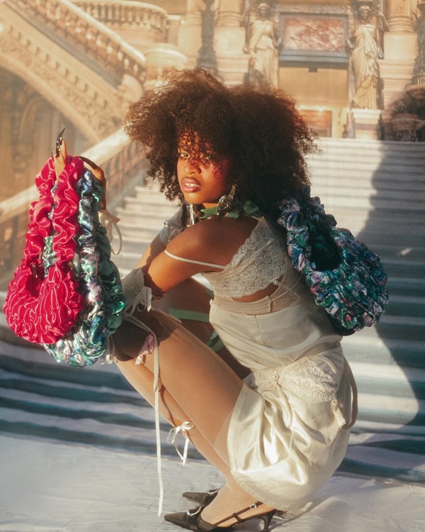 Mulher negra e jovem, de cabelo cacheado, posando para foto em uma escadaria de mármore. Ela usa uma camisola branca de seda e segura bolsas pequenas feitas em seda franzida nas cores azul, cinza e rosa