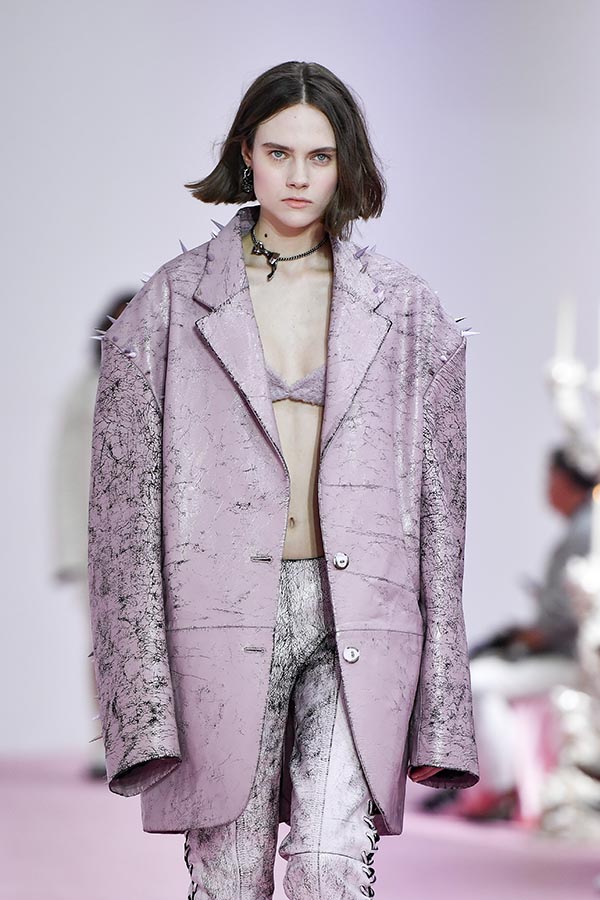 Uma modelo caminha na passarela durante o desfile de moda da Acne Studio, coleção de Primavera/Verão 2023, como parte da Paris Fashion Week em 28 de setembro de 2022. A modelo é uma mulher branca de cabelo liso curto e usa um blazer grande cinza metalizado. 