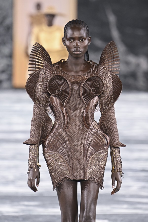 Uma modelo caminha na passarela durante o desfile de moda da Balmain, coleção de Primavera/Verão 2023, como parte da Paris Fashion Week em setembro de 2022. A modelo é uma mulher negra e jovem, de cabelo trançado longo, e usa um vestido marrom com mangas e ombreiras esculturais.