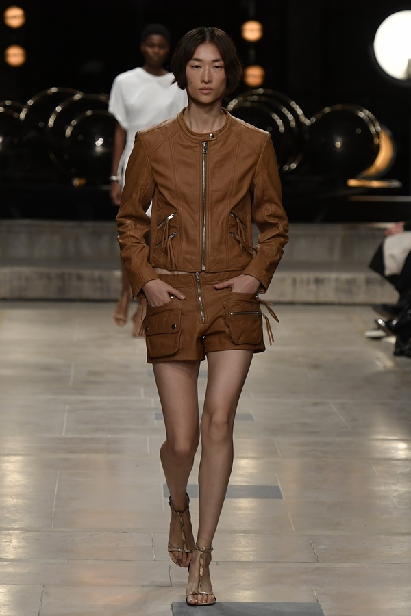 Uma modelo caminha na passarela durante o desfile de moda da marca Isabel Marant, coleção de Primavera/Verão 2023, como parte da Paris Fashion Week em setembro de 2022 em Paris, França. A modelo é uma mulher branca e jovem, de cabelos lisos curtos, e usa um casaco e um short de couro marrom.