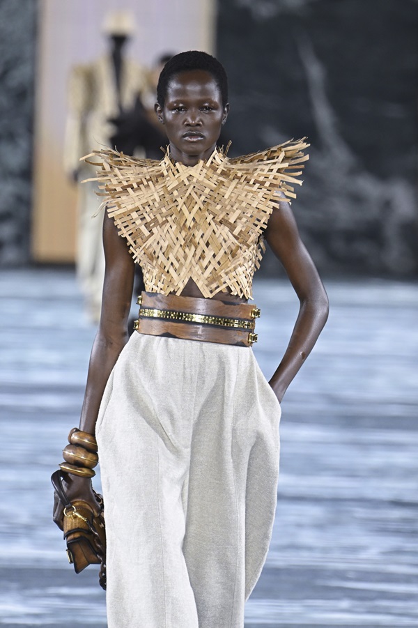 Modelo negra e jovem, de cabelo raspado crespo, andando na passarela do desfile da marca Balmain. Ela usa um top de palha, uma calça branca e um cinto de madeira.