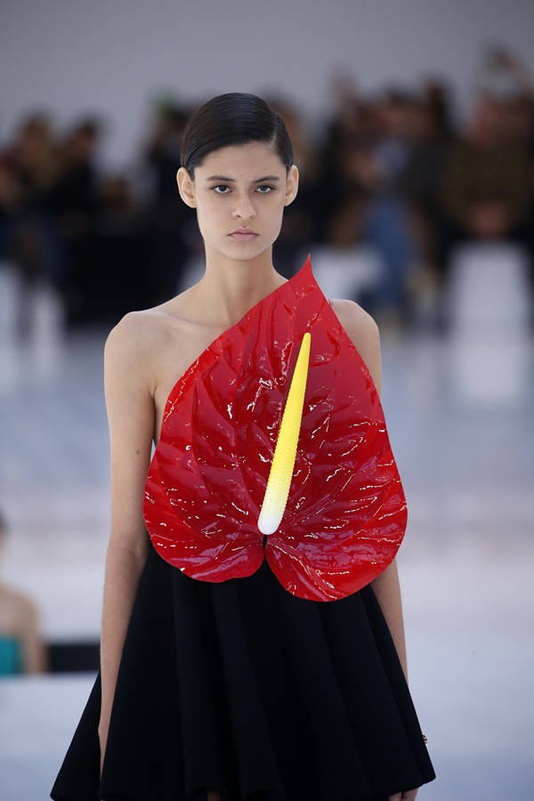 Modelo morena e jovem, de cabelo liso castanho, desfilando na apresentação da marca Loewe, em Paris. Ela usa um vestido com uma flor grande bordada na frente.