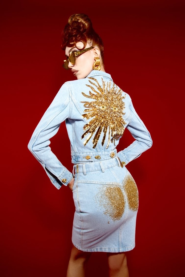 Mulher branca e jovem, de cabelo ruivo, posando para foto em um fundo vermelho. Ela usa um óculos de sol dourado, jaqueta e saia jeans com bordados dourados