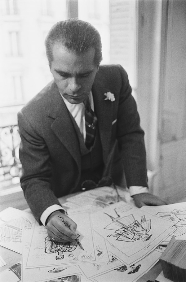 O estilista Karl Lagerfeld, em 1983, desenhando em seu estúdio criações de roupa para a marca Chloé. Ele usa um terno cinza, camisa branca e gravata xadrez.