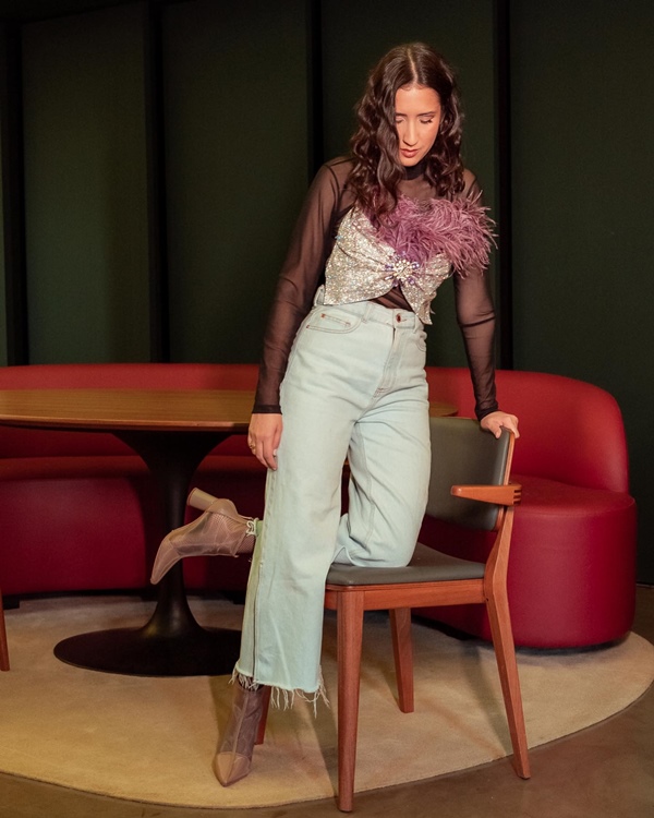 Mulher branca e jovem, de cabelo ondulado preto, posando para foto em um restaurante. Ela usa um top preto transparente de manga e, por cima, um curto prata de strass; calça jeans e uma bota bege