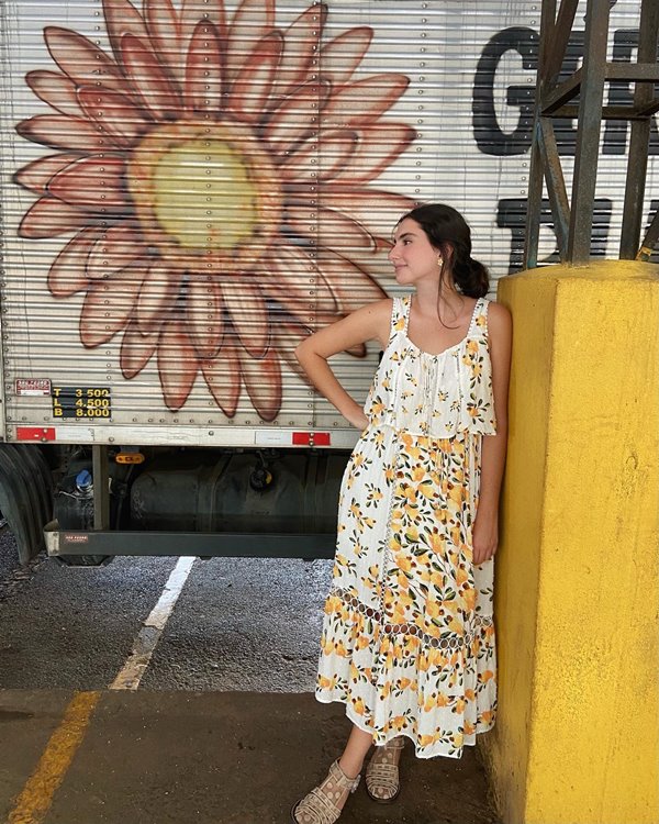 Mulher branca e jovem, de cabelo liso castanho, posando para foto em um estacionamento. Usa um vestido branco com estampa de flores amarelas