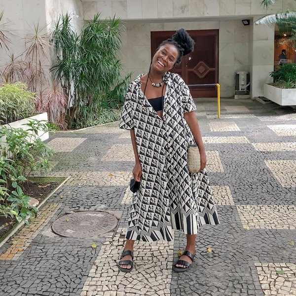 Mulher negra e jovem, de cabelo trançado amarrado, posando para foto na frente de um prédio. Usa um vestido preto e branco listrado