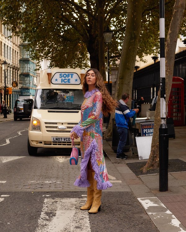 Mulher jovem e branca, de cabelo longo e ondulado, posando para foto nas ruas de Londres. Ela usa um vestido estampado e botas de couro marrom