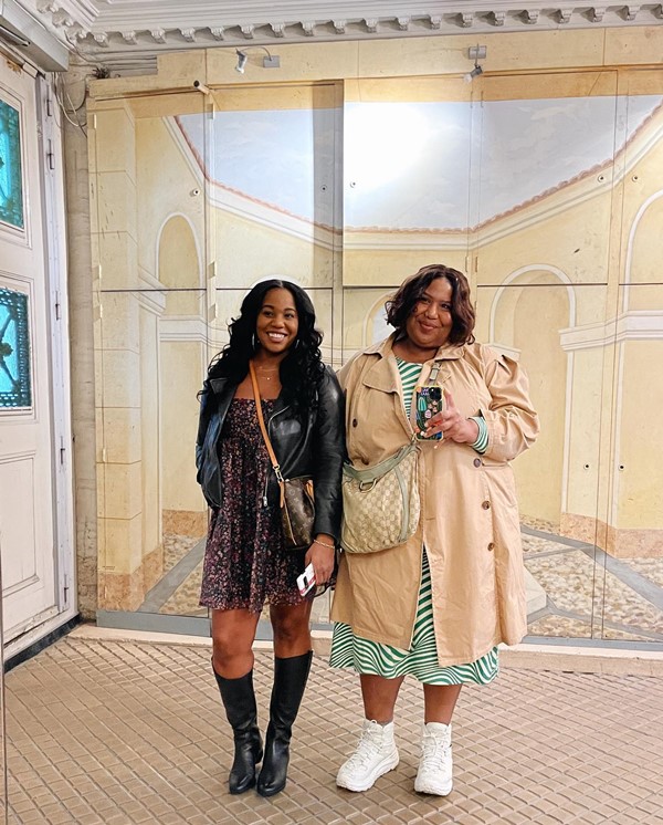 Duas mulheres jovens e negras posando para foto no espelho de uma galeria. A primeira usa um vestido florido escuro, jaqueta e bota de couro; e a segunda um vestido listrado verde e branco, sobretudo bege e tênis branco