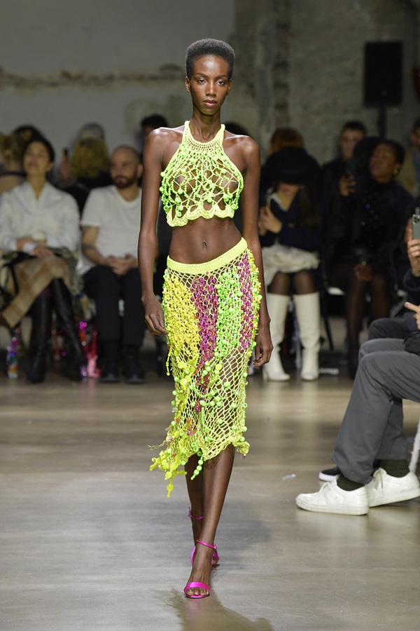 Modelo negra e jovem, de cabelo crespo curto, desfila durante a apresentação da marca Germanier, na temporada de Primavera/Verão 2023, como parte da Semana de Moda em 03 de outubro de 2022 em Paris, França. Ela usa um top curto de corda verde e uma saia, também de corda, verde e rosa.