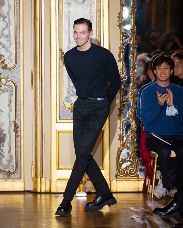 O estilista Kevin Germanier, um homem jovem, branco, de cabelo curto castanho, na fila final do próprio desfile com roupas todas pretas