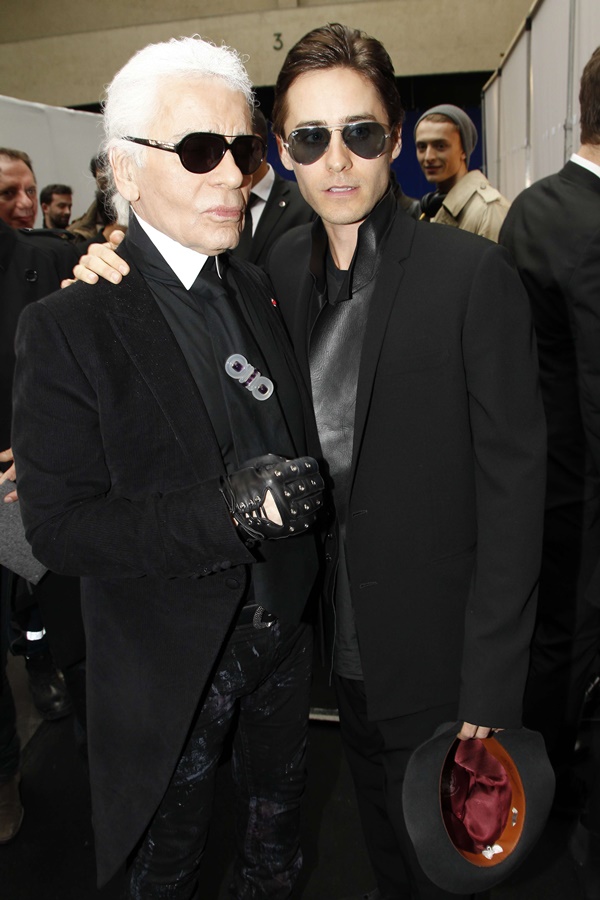 O estilista Karl Lagerfeld, um homem branco, idoso e de cabelos brancos e lisos comprimentando o ator Jared Leto, um homem branco de cabelo liso castanho. A foto é de 2012 em um desfile da linha masculina da marca Dior Homme.