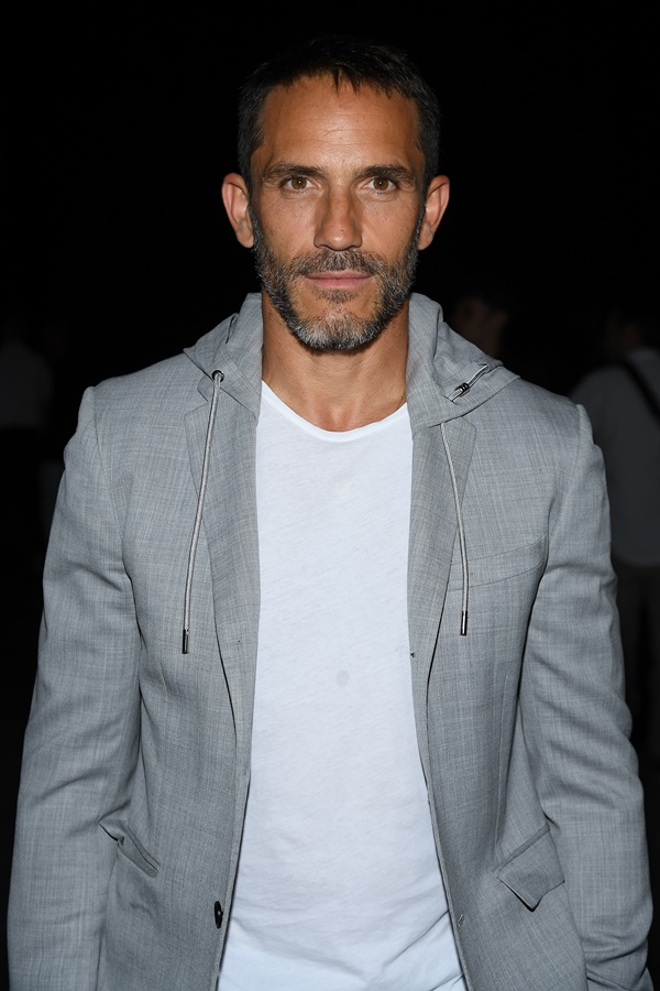 Sébastien Jondeau, um homem branco, de meia idade e cabelo e barba grisalha, posa para foto no desfile da Celine em 2019. Ele usa uma camiseta branca e um casaco cinza