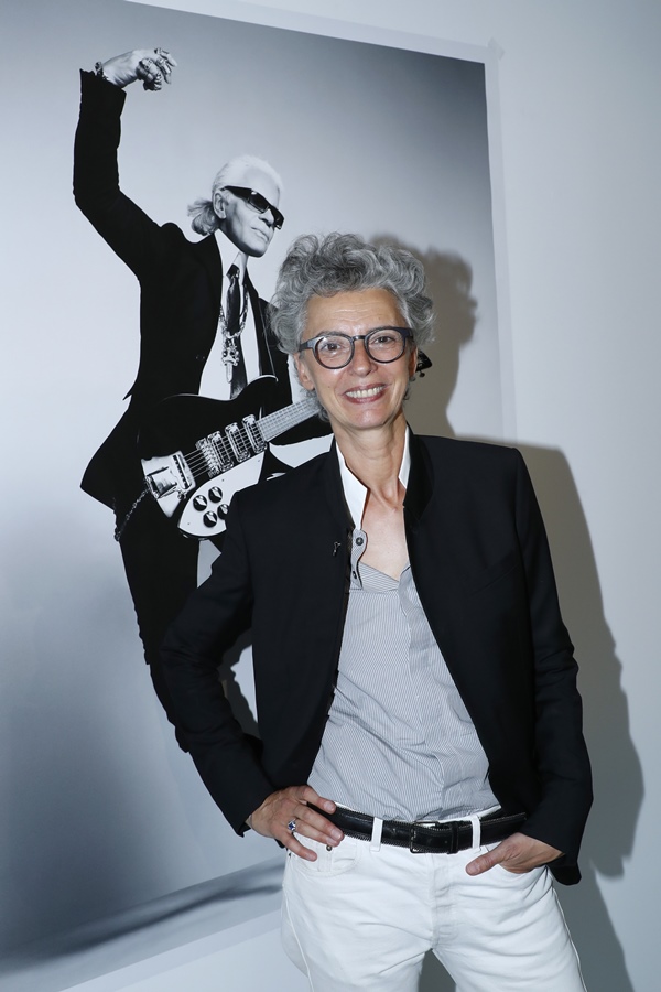 Caroline Lebar, uma mulher de meia idade, branca e de cabelos ondulados grisalhos, posa para foto em frente a um quadro com a foto do estilista Karl Lagerfeld