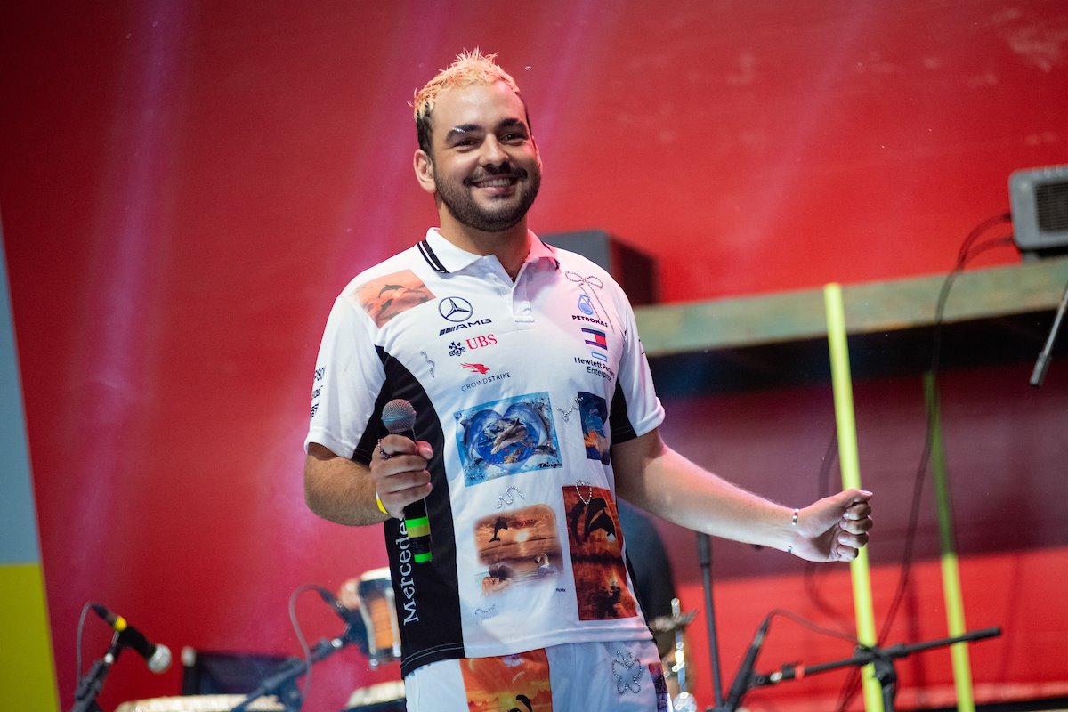 O cantor Totô de Babalong, um homem branco, jovem, de barba castanha e cabelo curto loiro, cantando no palco do evento Metrópoles Fashion & Design