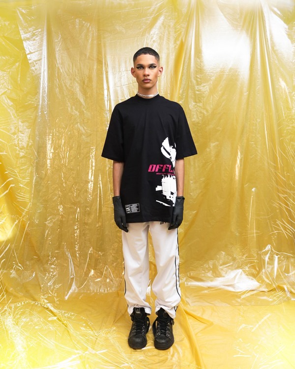 Ensaio fotográfico da marca brasiliense Off Shadow feito em um estúdio com um fundo amarelo plastificado. Na foto é possível ver um modelo homem, negro e jovem, com cabelo curto raspado, usando uma camiseta preta, calça larga branca e tênis e luvas de couro preto.