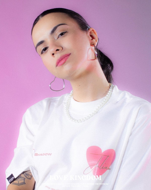 Ensaio fotográfico da marca brasiliense Off Shadow feito em um estúdio com um fundo lilás liso. Na foto é possível ver um mulher branca e jovem, de cabelos lisos castanhos, usando uma camiseta branca, uma corrente de pérola e um brinco de argola prata.