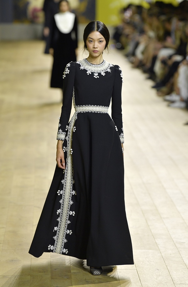 Uma modelo caminha na passarela durante o desfile Christian Dior Haute Couture Fall Winter 2022 2023 como parte da Paris Fashion Week em 04 de julho de 2022 em Paris, França. Ela é uma mulher branca, de cabelo liso preto e longo, com traços asiáticos, e usa um vestido preto com detalhes de flores bordadas.