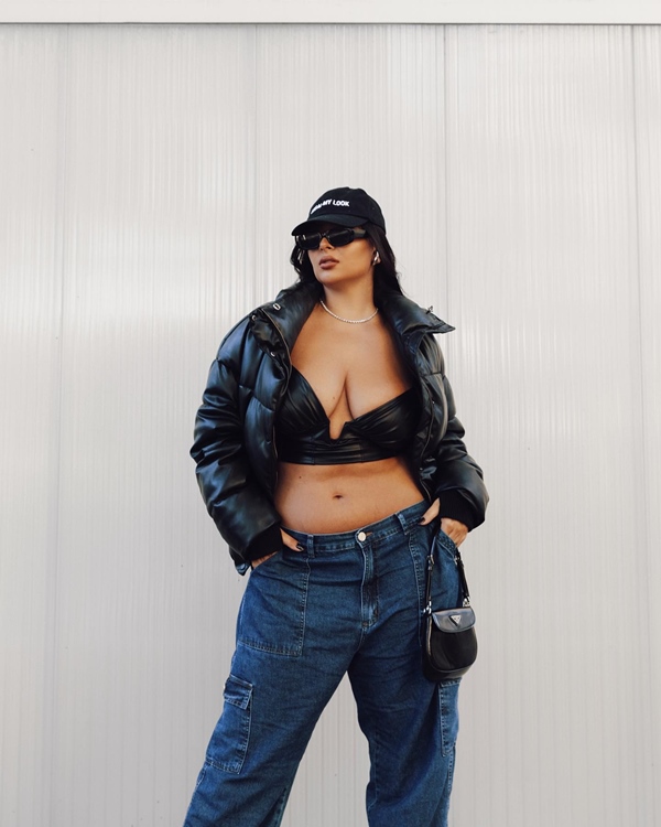 Mulher jovem e branca, de cabelo longo liso, posando para foto em frente a um mural branco. Ela usa um top curto preto, calça jeans folgada, jaqueta de couro, boné preto e óculos escuros.