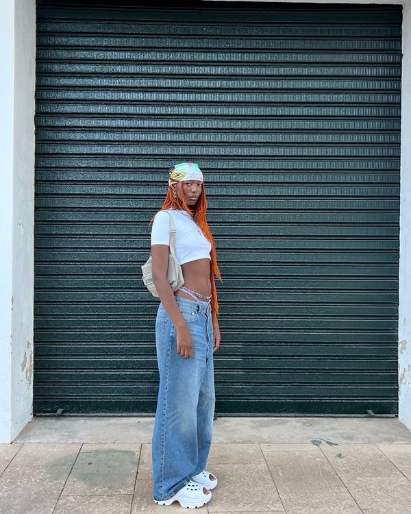 Mulher jovem e negra, de cabelo trançado ruivo e longo, posando para foto na rua, em frente a um portão de metal. Ela usa um top cropped branco, calça jeans folgada e um lenço amarrado na cabeça