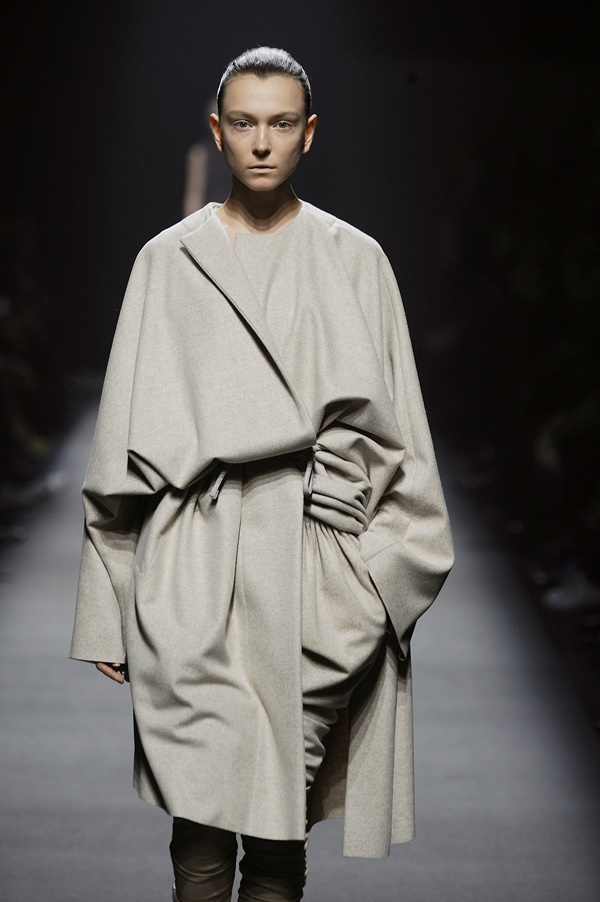 Uma modelo jovem e branca, com o cabelo liso amarrado em um coque, desfila com a coleção Outono/Inverno 2008/2009 da marca Haider Ackermann durante a Paris Fashion Week no dia 28 de fevereiro de 2008 em Paris, França. Ela usa um casaco de inverno bege com cinto amarrado em um laço na cintura - Metrópoles