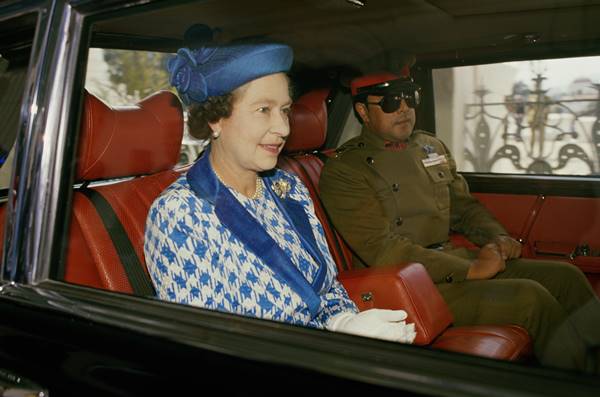 Rainha Elizabeth II no Nepal usando terno com estampa pied de poule branca e azul. Ela está em um carro ao lado de uma autoridade do país com traje militar - Metrópoles