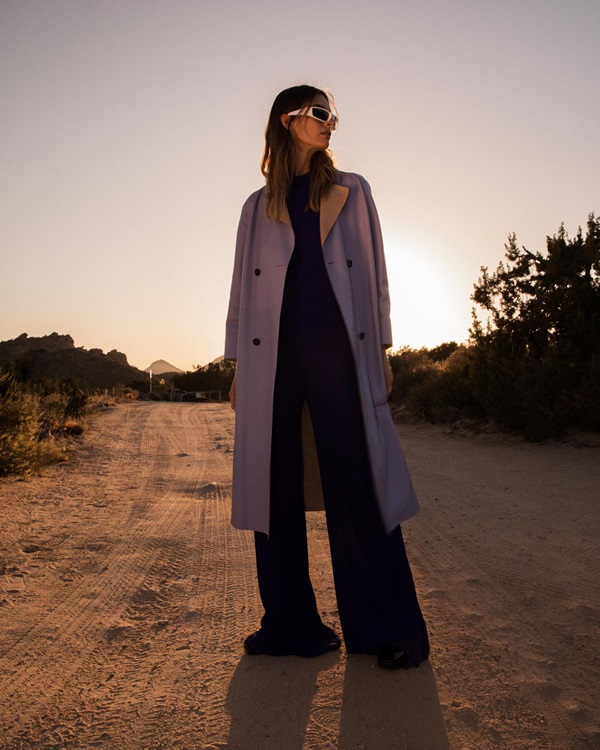 Foto de campanha da marca Maison Ullens, onde posa uma modelo mulher jovem, de pele clara e cabelo liso castanho, em uma estrada sem asfalto. Ela usa uma blusa preta, uma calça larga também preta e um casaco bege estilo trench coat - Metrópoles