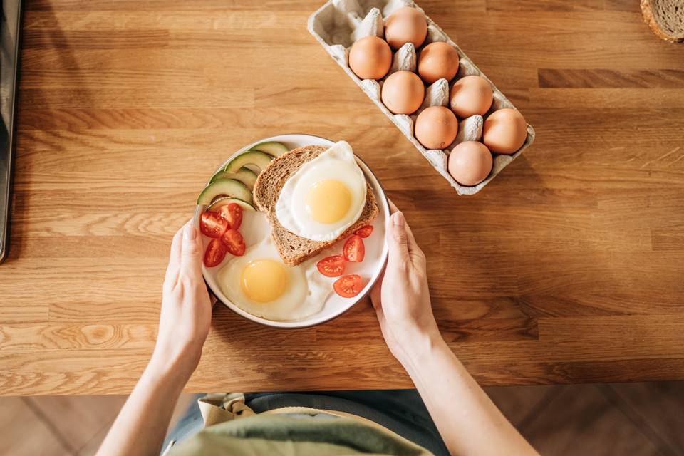 Fotografia mostra mãos segurando um prato de comida cheia de proteínas e uma caixa de ovos em cima da mesa - Metrópoles