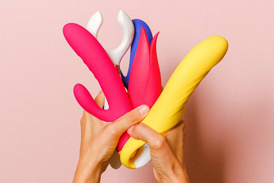 Pessoa segura diversos sex toys coloridos sob fundo rosa - Metrópoles