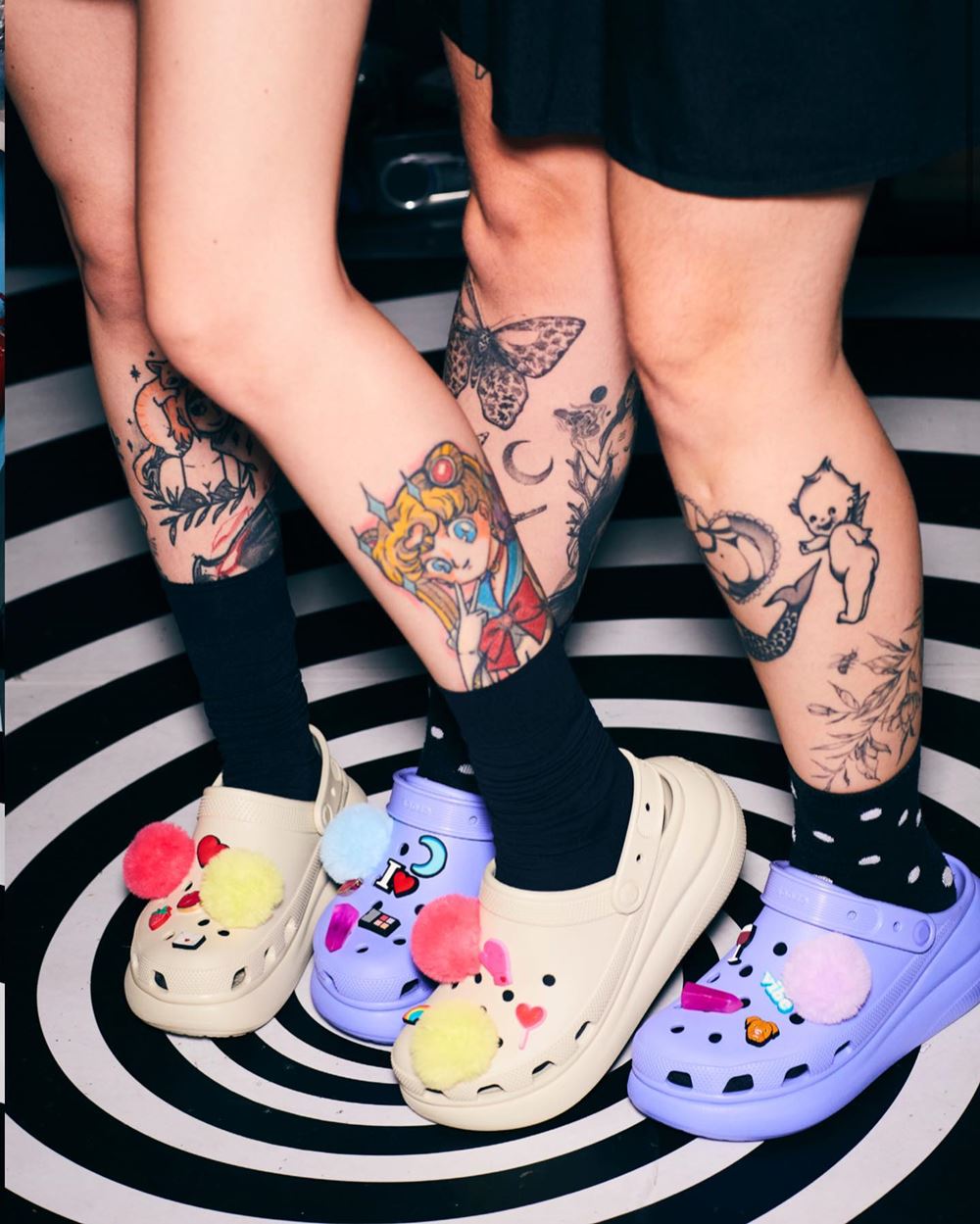 Duas mulheres jovens e de cabelo liso colorido posando para foto em uma campanha da marca Crocs. Elas usam roupas pretas, possuem tatuagens e usam meias, também pretas, com os sapatos da marca coloridos: um rosa e um lilás - Metrópoles