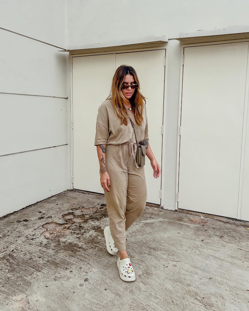 Ju Bandeira, influencer de Brasília, uma mulher morena de cabelo liso loiro, posando para foto em frente ao portão de casa. Ela usa uma camiseta bege, uma calça de moletom da mesma cor e um sapato crocs bege - Metrópoles