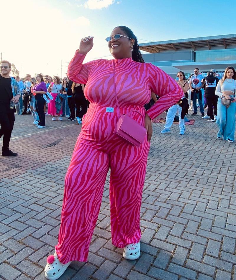 Tiane Felix, influenciadora, uma mulher negra de cabelo liso preto, posando para foto no festival Rock in Rio. Ela usa um conjunto de calça e blusa de manga longa rosa, com estampa de listras vermelhas, óculos escuros, uma bolsa rosa e uma sandália crocs bege - Metrópoles