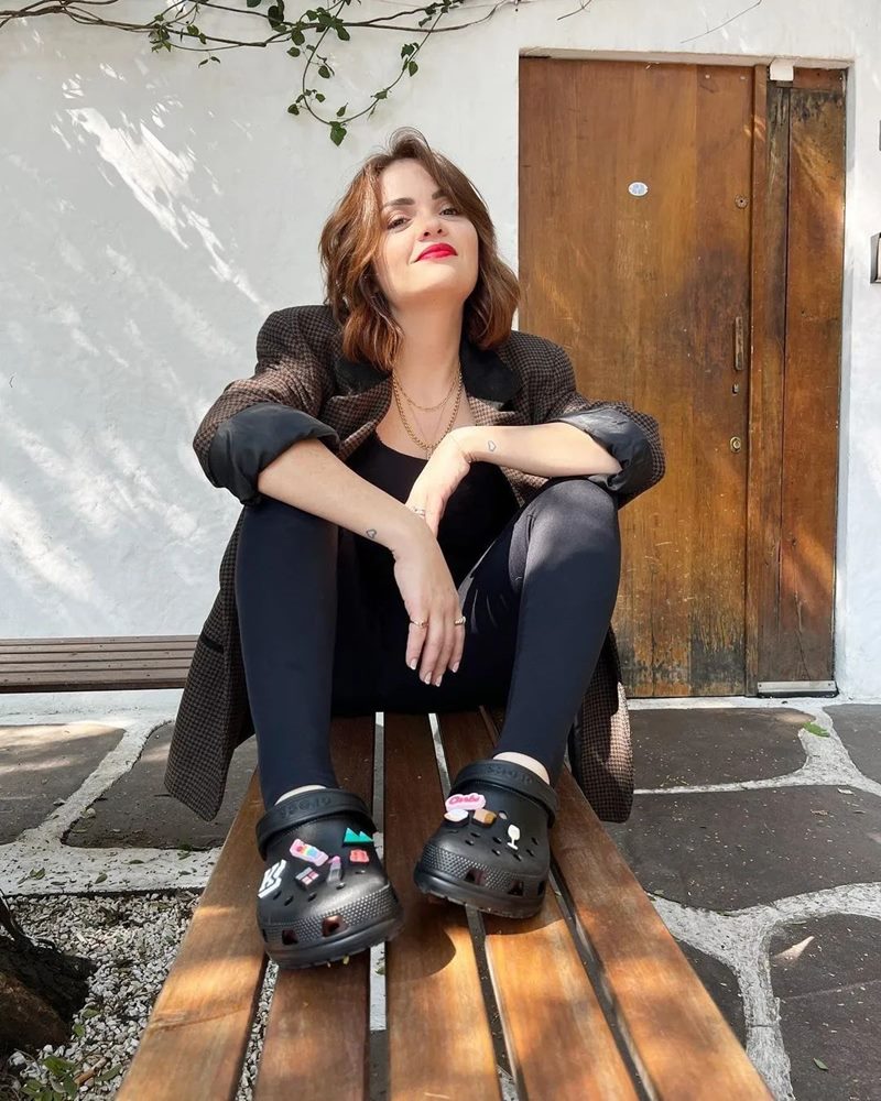 Karol Pinheiro, influenciadora, uma mulher branca de cabelo ruivo curto e onduldo, posando para foto em frente ao portão de casa. Ela usa camiseta, calça legging, blazer e um sapato crocs, tudo na cor preta - Metrópoles